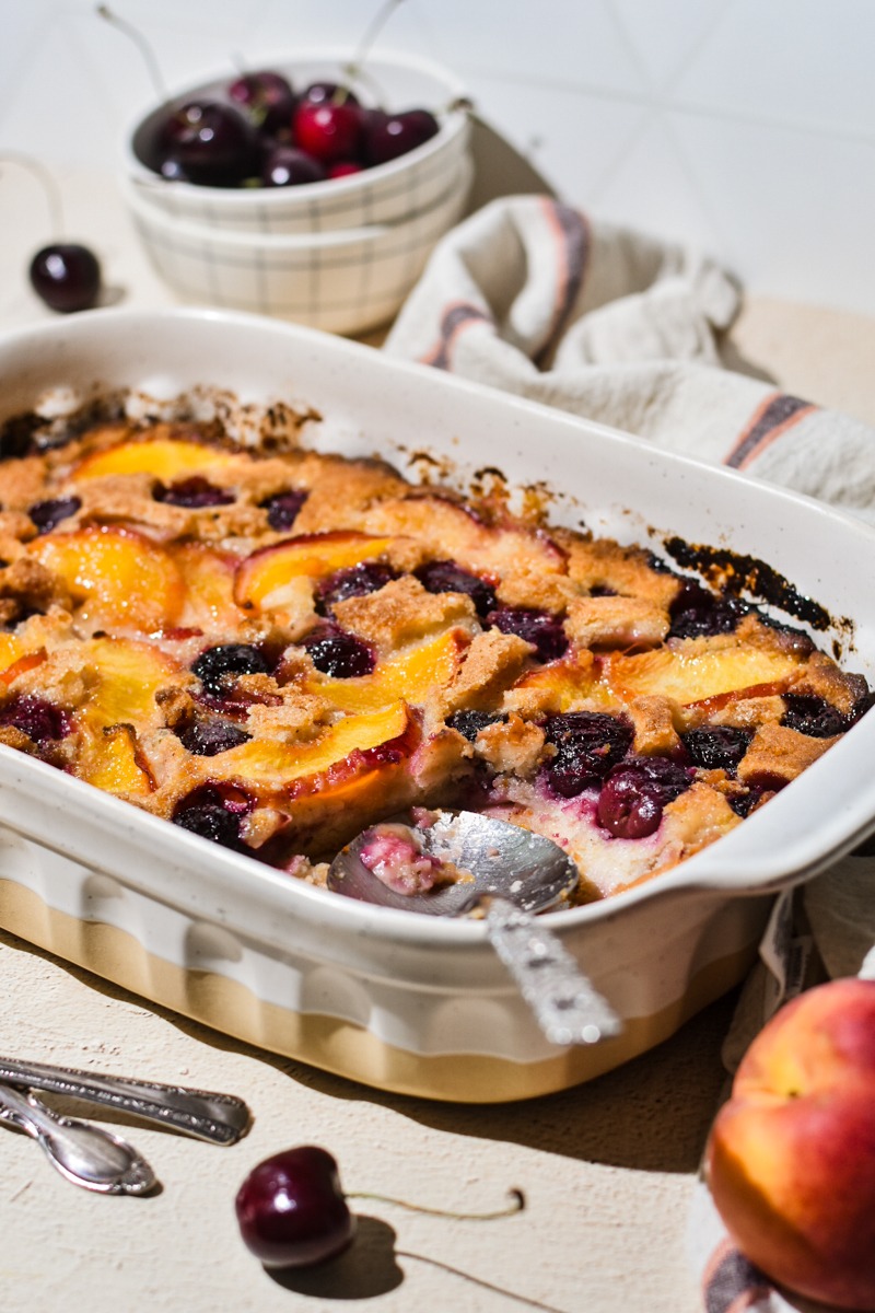 Old-fashioned peach cobbler with cherries in a white baking dish.