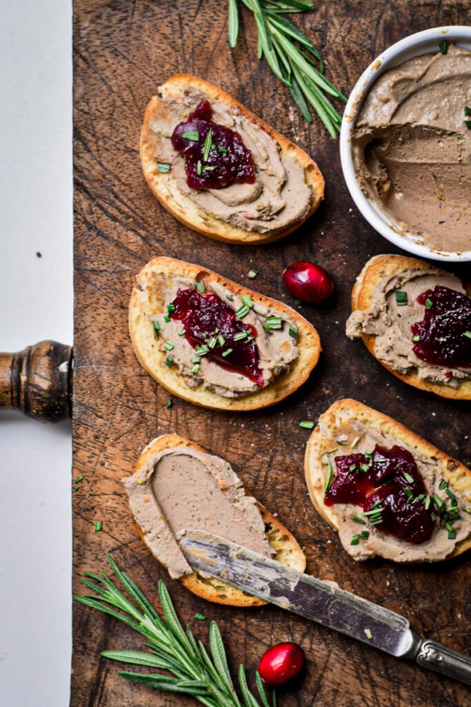 Crostinis in a wooden board with chicken liver pate on top and a dollop of jam on top of that.