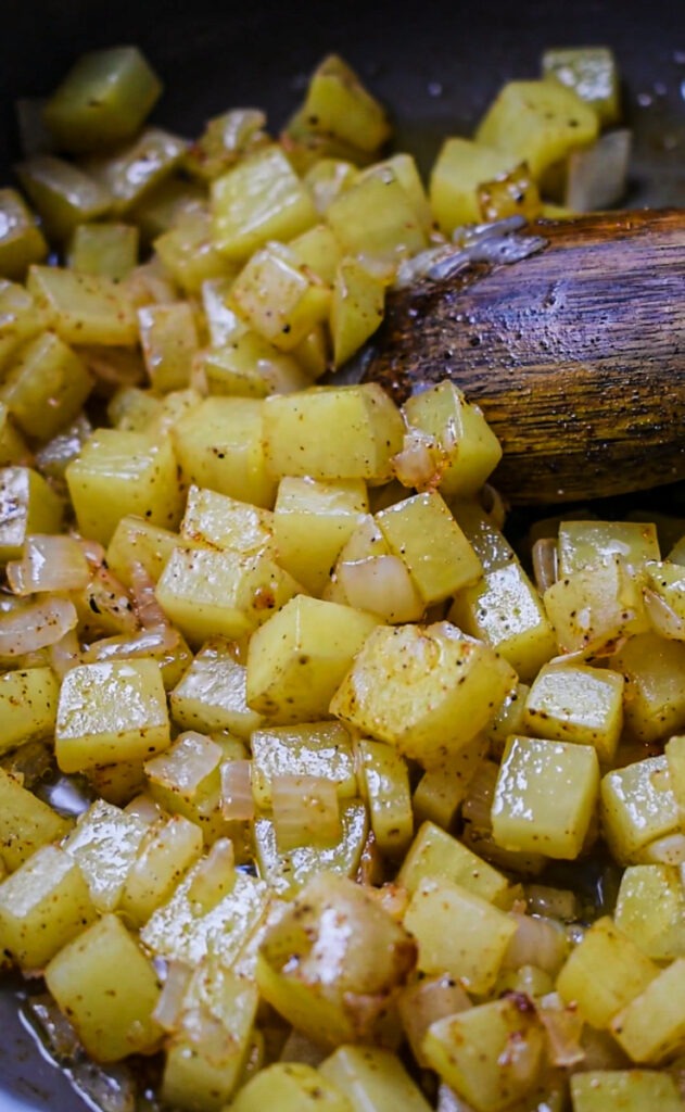 Diced potatoes cooking in a skillet with a wooden spoon stirring them.
