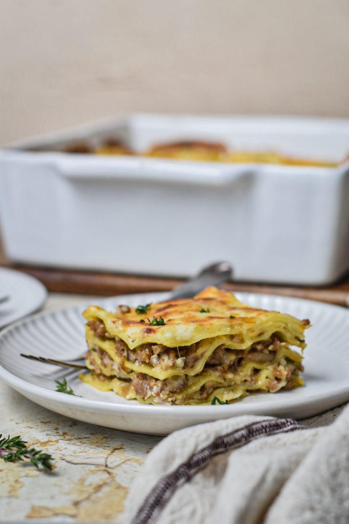 Slice of mushroom and sausage lasagna on a white plate with more lasagna in the background.