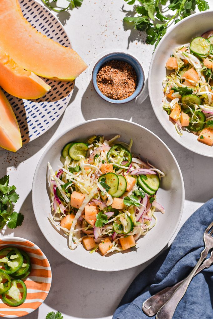 Two bowls of cantaloup cabbage slaw surrounded by other ingredients like cantaloupe slices and a small bowl of sliced jalapeño.