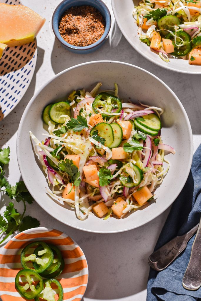 Spicy melon slaw served in a white speckled bowl surrounded by other ingredients.