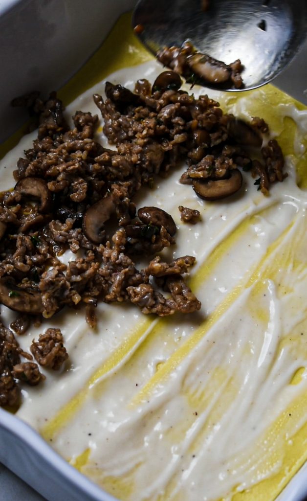 Sausage and mushroom filling being layered on top of the bechamel sauce.