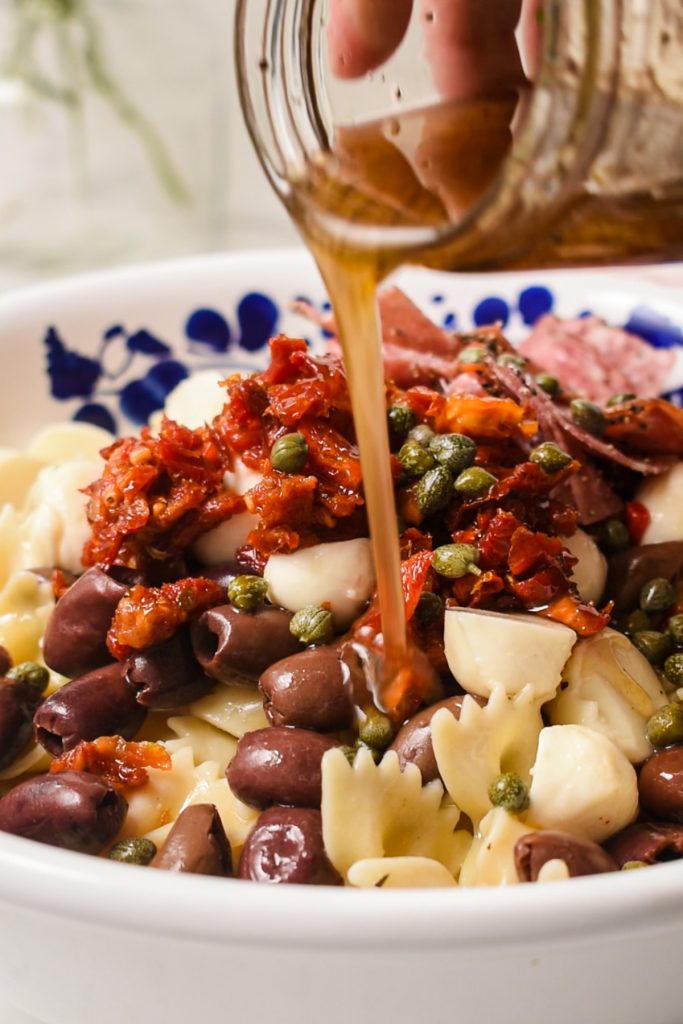 Hand pouring the homemade dressing on top of the pasta salad in a bowl.