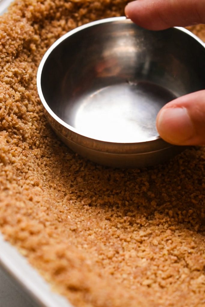 Hand using a measuring cup to press and shape the graham cracker crust into the pie dish.