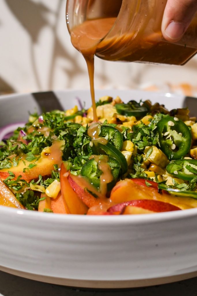 Hand pouring the homemade dressing from a jar onto the summer corn peach coleslaw.