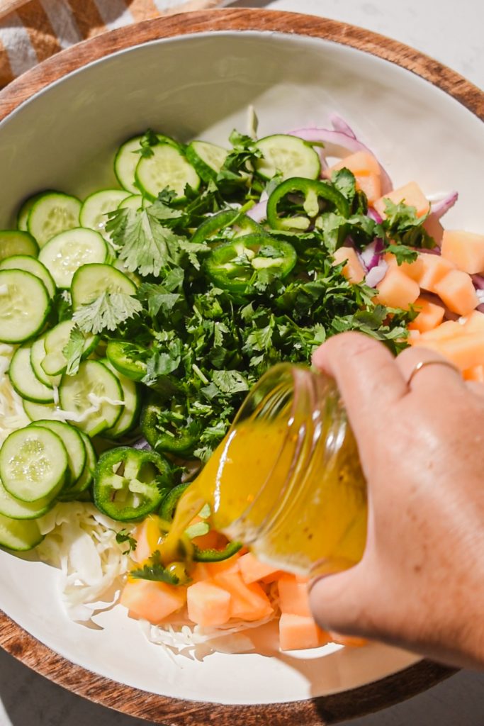 Hand pouring the dressing over the slaw ingredients in a bowl.