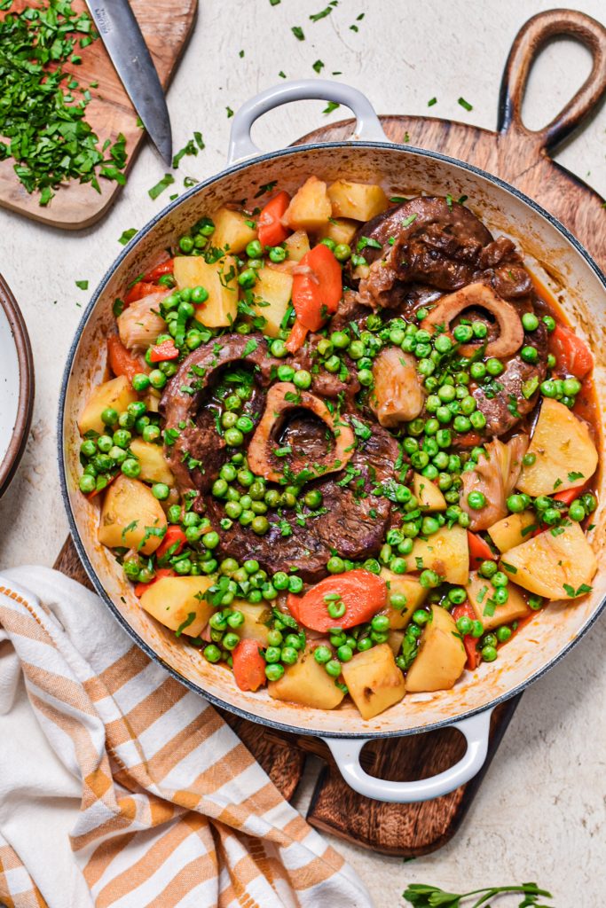 Overhead shot of spring stew osso buco in a Dutch oven on a round wooden cutting board.