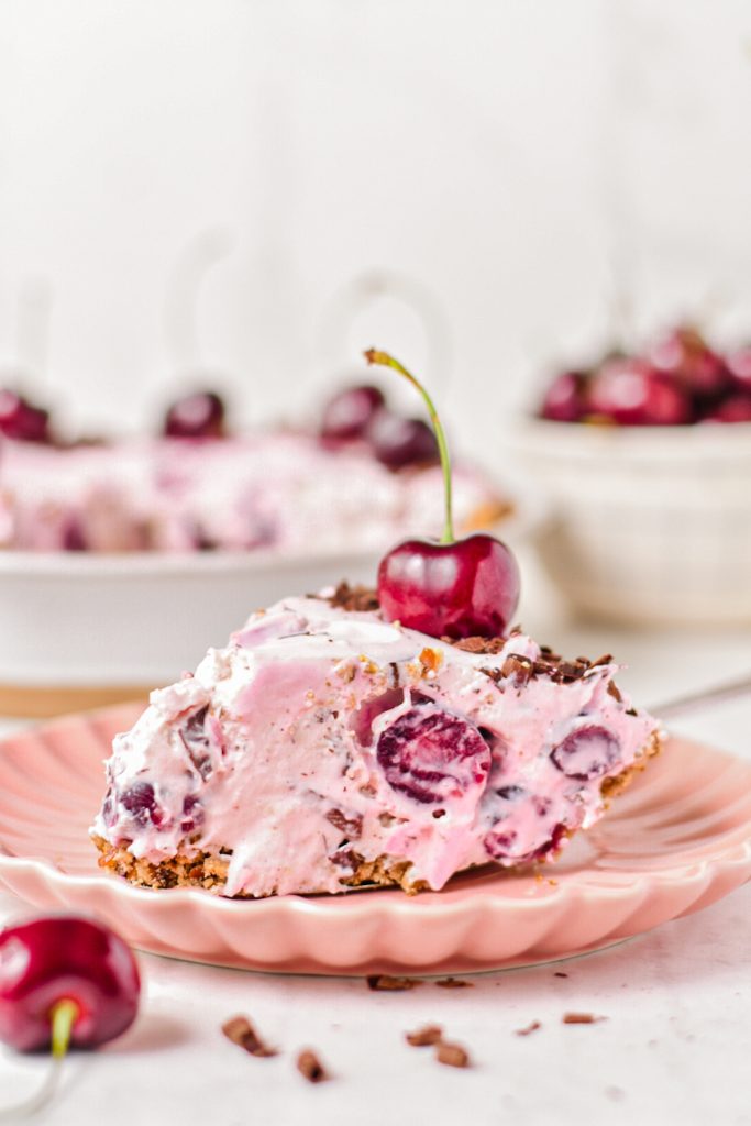 Slice of cherry chocolate pie on a pink plate with a fresh cherry on top and the pie in the background.