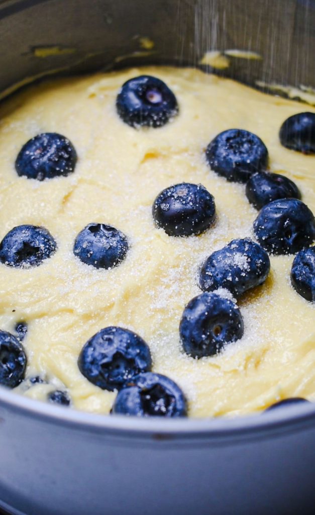 Cake batter and fresh blueberries in a cake pan with a hand sprinkling sugar on top.