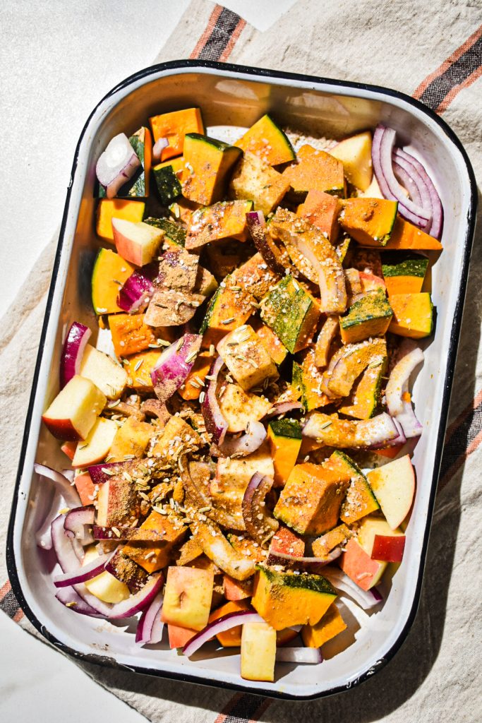 Raw squash, apple, and red onion being seasoned in a baking dish.