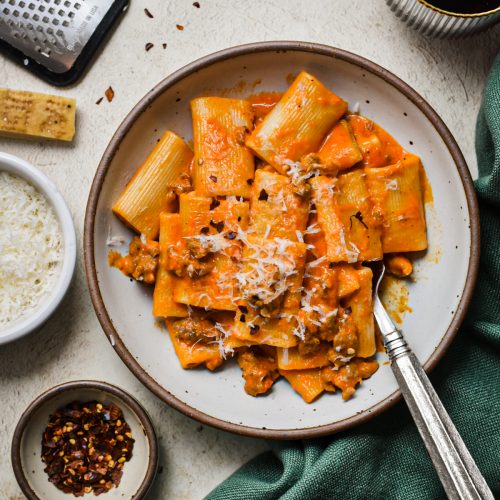 Italian sausage and red pepper pasta served in a bowl next to a small serving of chili flakes and parmesan.