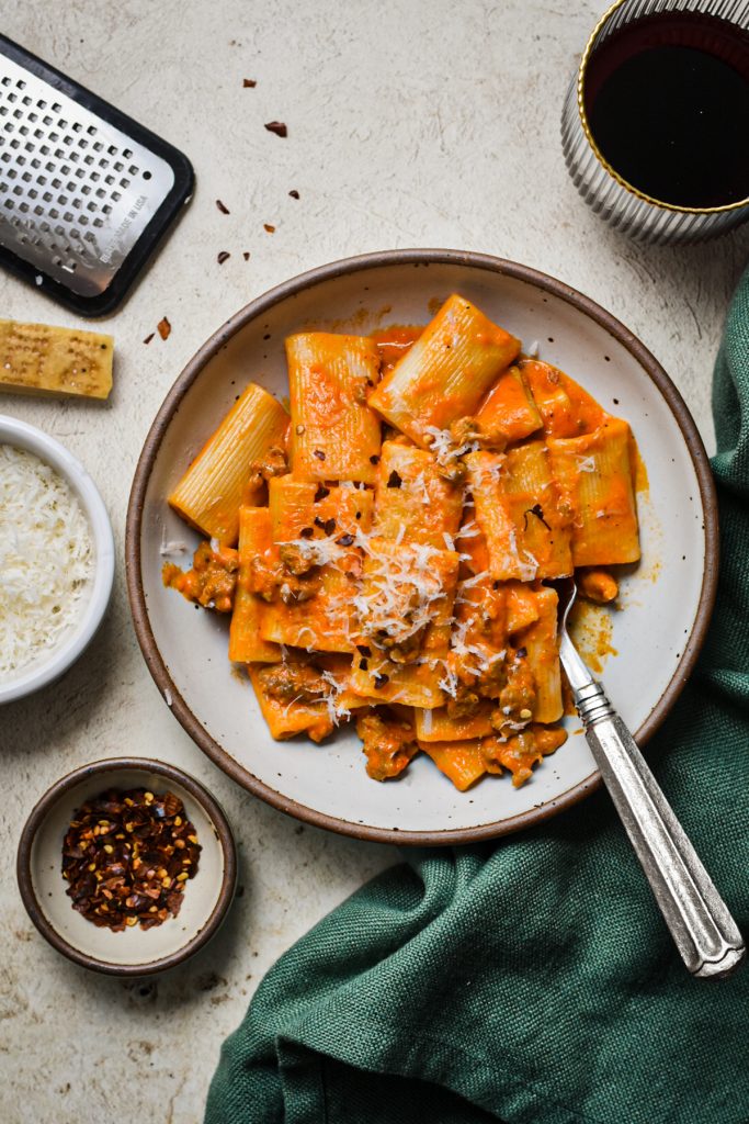 Italian sausage and red pepper pasta served in a bowl next to a small serving of chili flakes and parmesan.