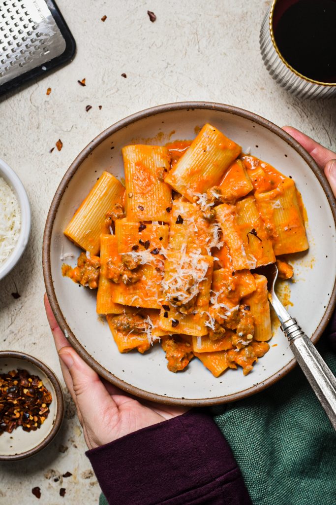 Two hands holding onto a bowl of Italian sausage and red pepper pasta.