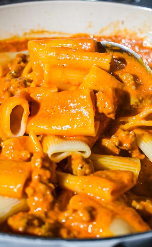 Cooked pasta being combined with the sauce and sausage in a skillet.
