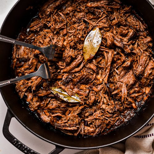 Beef barbacoa being shredded in a cast iron pan.