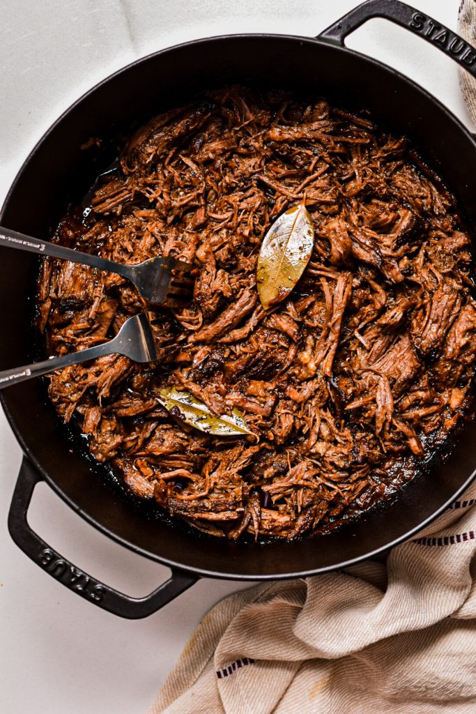 Beef barbacoa being shredded in a cast iron pan.