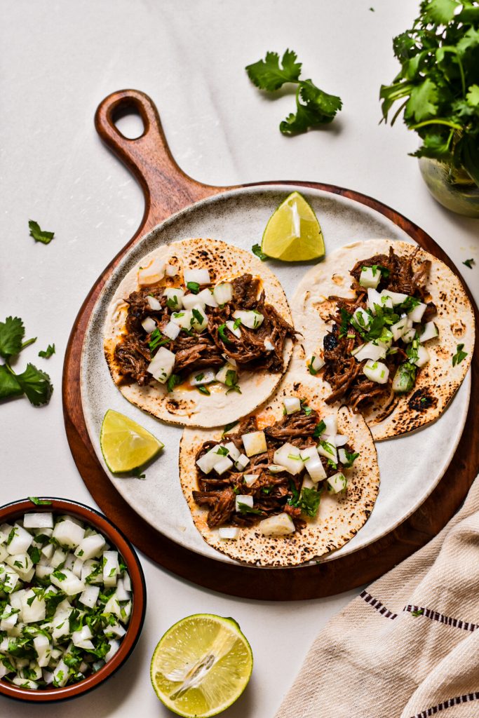 Three beef barbacoa tacos on a serving plate with lime wedges.
