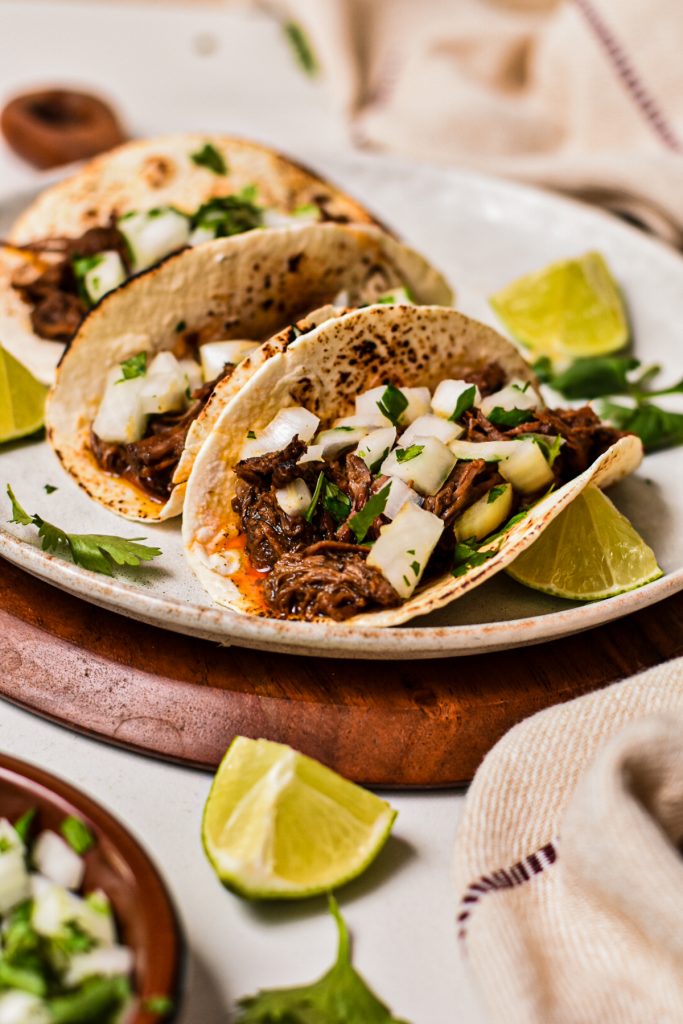 Three barbacoa tacos on a plate topped with onions and cilantro.