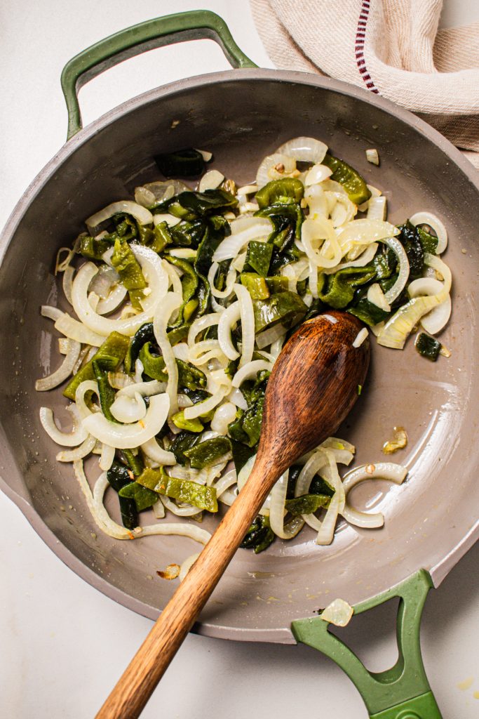 Onions and peppers cooking in a skillet with a wooden spoon.
