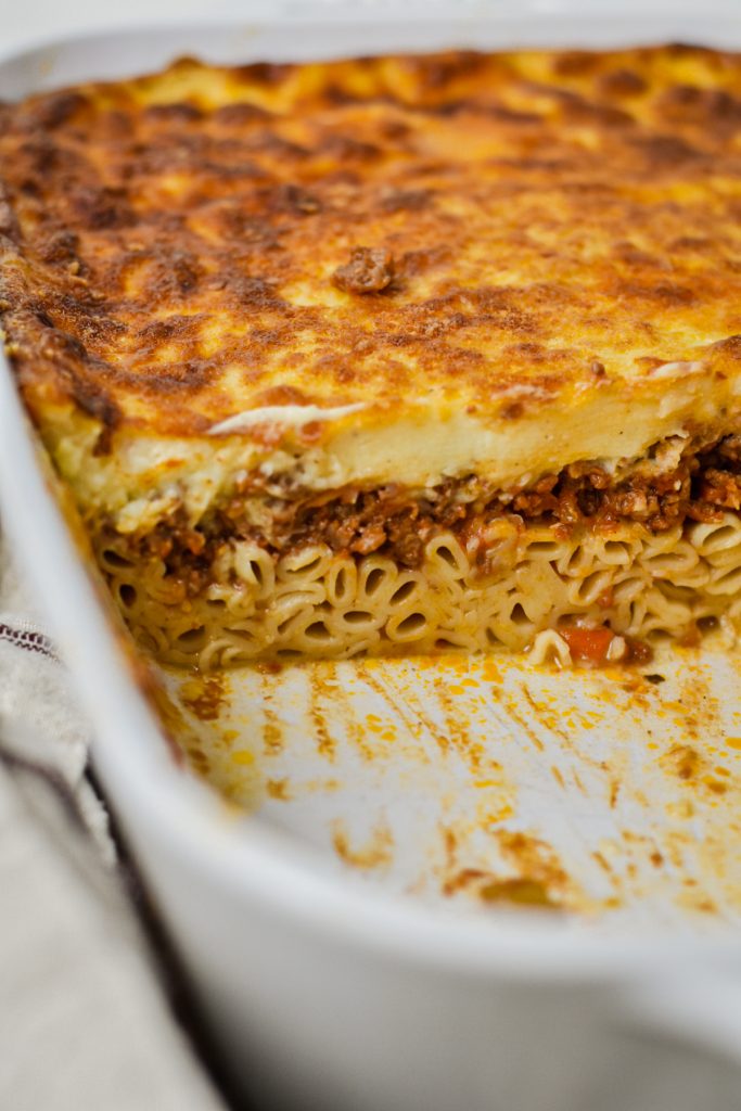 White baking dish of Greek pastitsio with a slice taken to show the layers of noodles, meat sauce and white sauce.