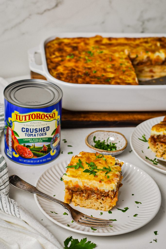 Slice of Greek Pastitsio on a plate next to a can of Tuttorosso crushed tomatoes with more pastitsio in the background.