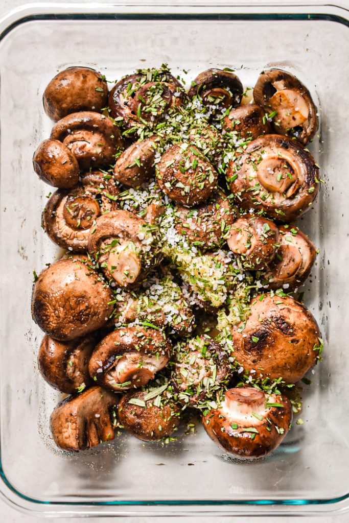 Cooked mushrooms in a glass container with seasonings like rosemary, salt and pepper on top.