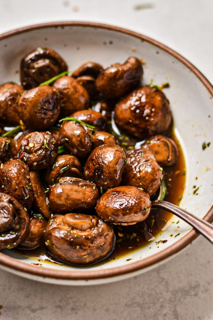 Balsamic marinated mushrooms topped with fresh rosemary in a bowl with a serving spoon.