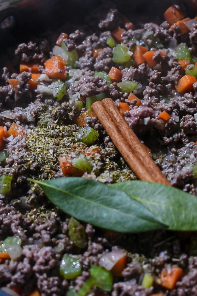 Ground beef in a skillet with veggies, seasonings, bay leaves and a cinnamon stick.