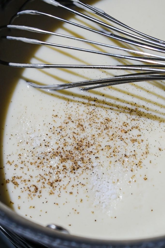 Whisking the bechamel sauce in a saucepan with salt and pepper being added.
