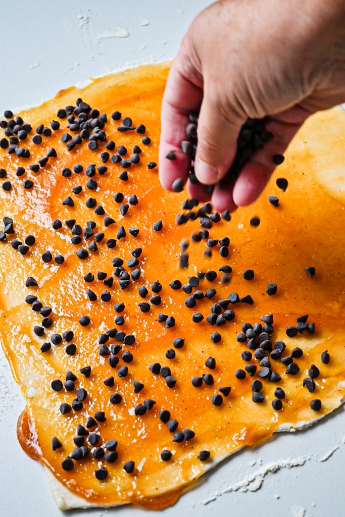 Sprinkling mini chocolate chips on top of the layer of pumpkin butter on a sheet of puff pastry.