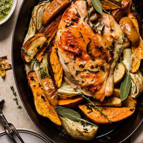 Maple roasted turkey breast in a roasting pan with roasted vegetables around it and fresh herbs on top.