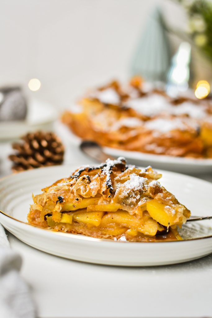 Slice of flaky apple tart on a plate with the rest of the tart in the background.