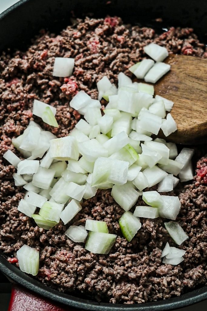 Ground beef in a skillet with chopped onions being added to it.