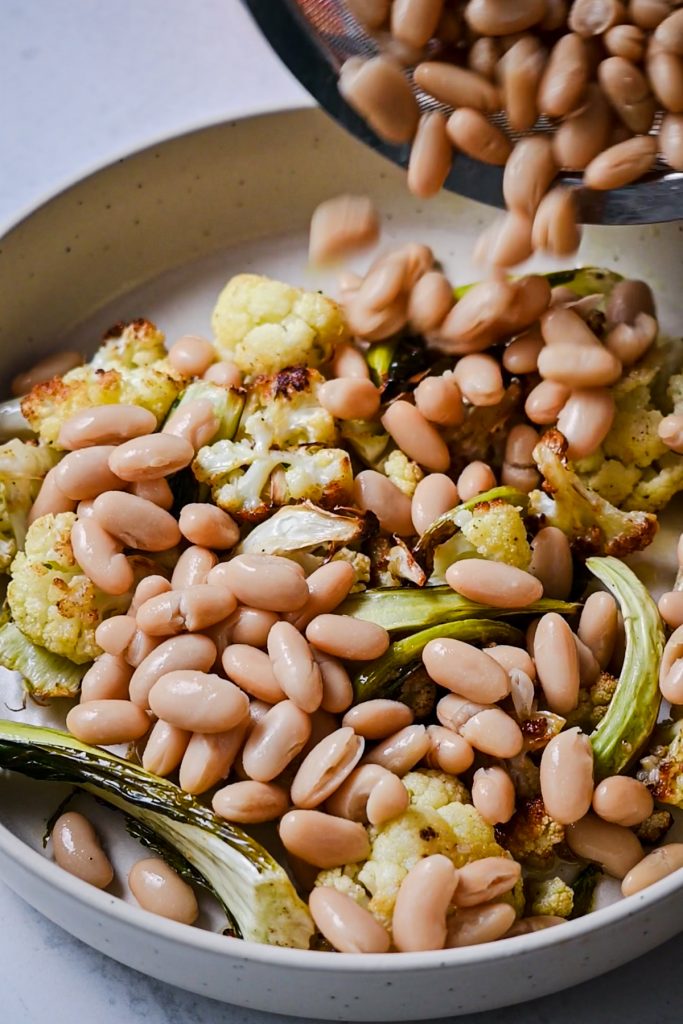 Pouring the rinsed beans into a bowl of roasted cauliflower.