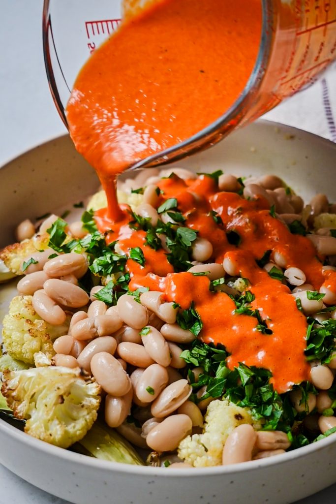 Drizzling the roasted red pepper sauce onto the warm cauliflower salad in a bowl.