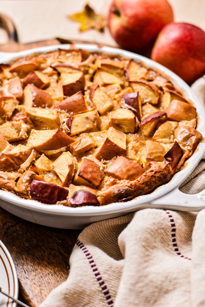Side view of an apple flaugnarde in a white pie dish with fresh apples in the background.