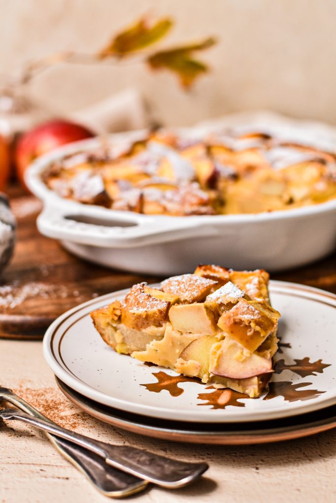 Slice of apple flaugnarde on a plate with the whole flaugnarde in the background.