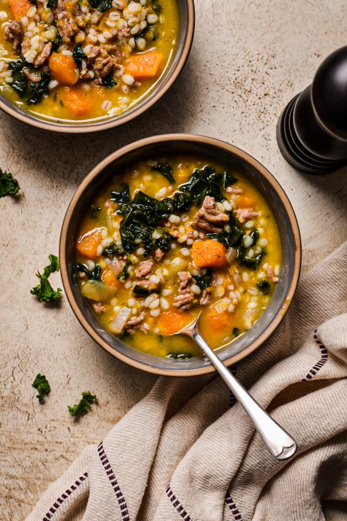 Overhead shot of a bowl of butternut squash, sausage, kale and barley soup with a spoon in the bowl.