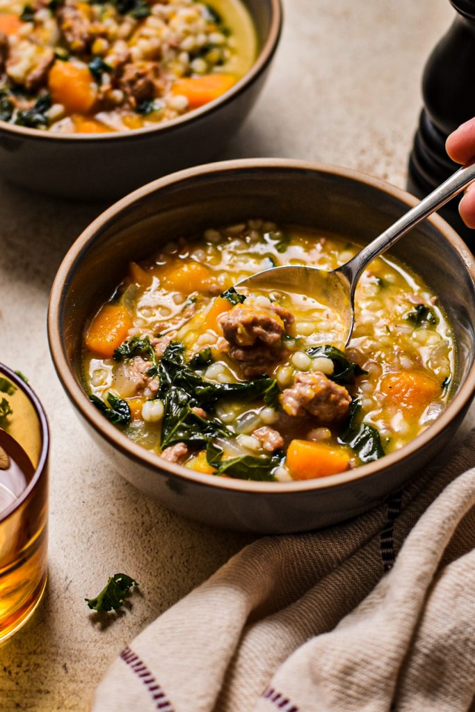 Butternut squash, sausage, kale and barley soup in a bowl with a hand taking a spoonful.