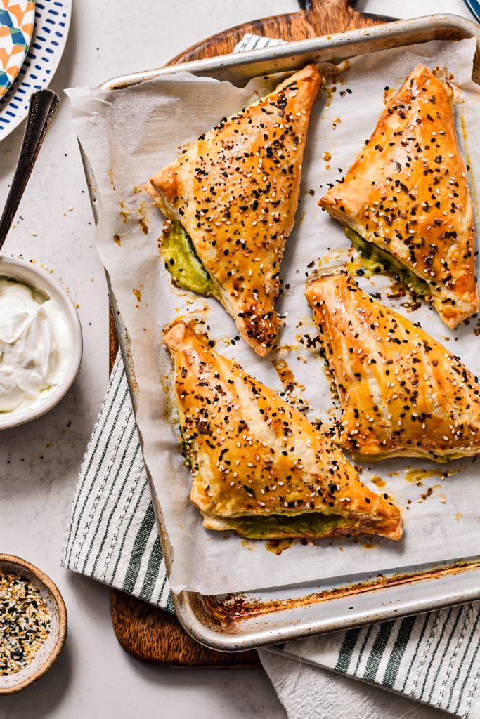 Four spanakopita turnovers on a sheet pan lined with parchment paper next to yogurt in a bowl.