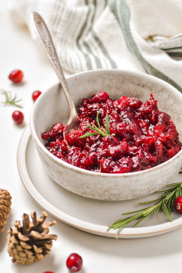 Roasted cranberry sauce in a serving bowl with a spoon and rosemary garnish.