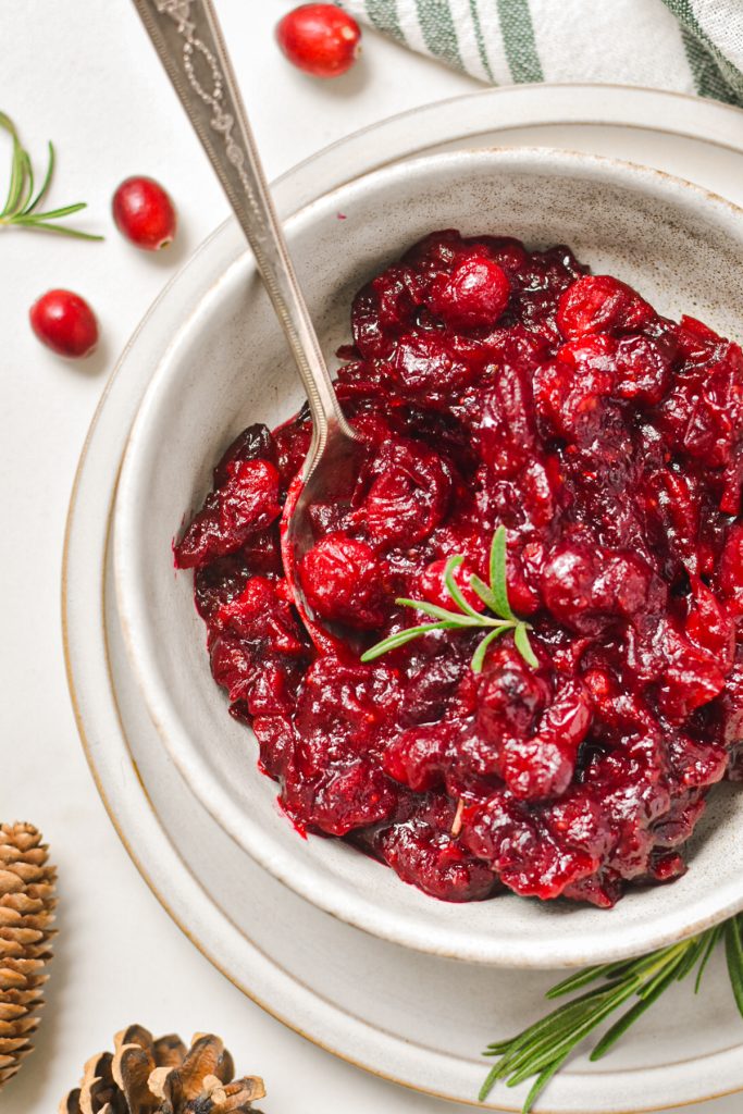 Bright red roasted cranberry sauce in a serving bowl with rosemary as garnish.