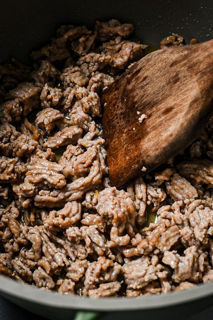Italian sausage being browned in the bottom of a large pot with a wooden spoon breaking it up.