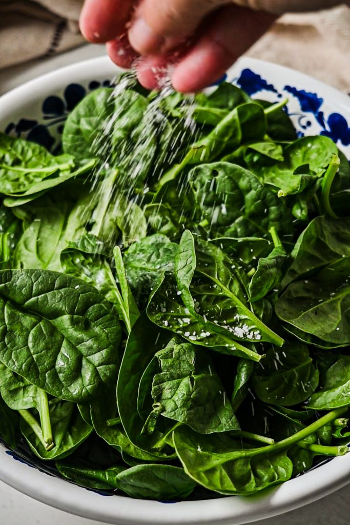 Sprinkling salt into a bowl of fresh spinach.