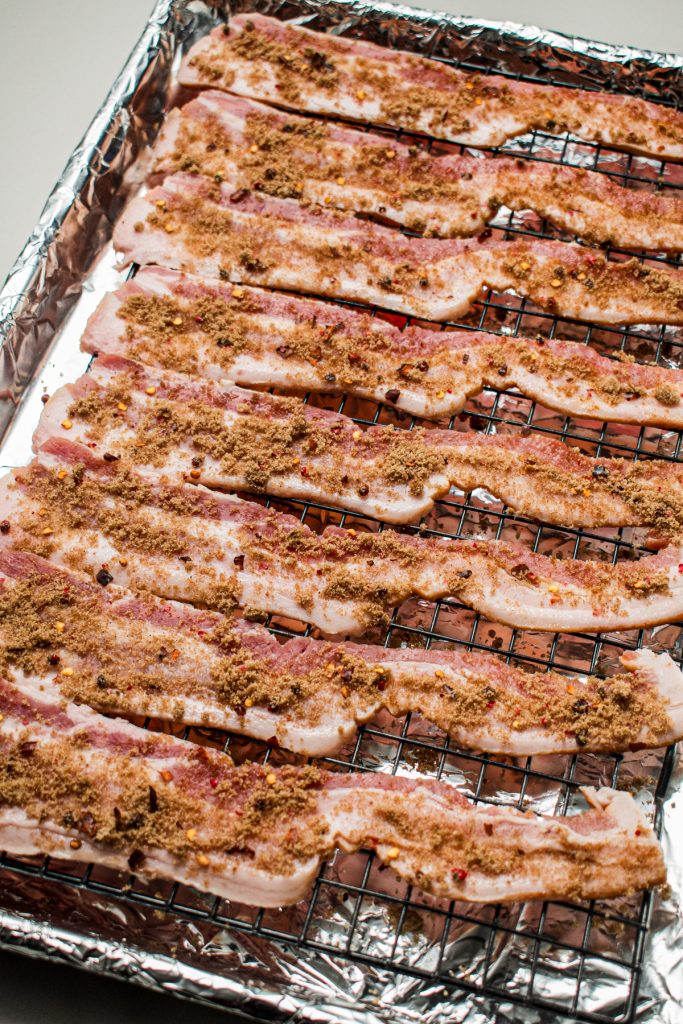 Raw bacon lined up on a baking sheet with the sugar and spice mixture sprinkled on top of it.