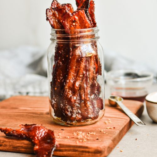 Candied bacon placed in a jar on top of a wooden board with one piece of bacon on the board.
