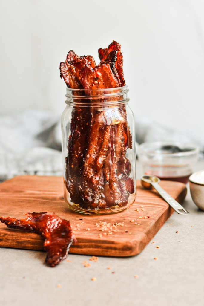 Candied bacon placed in a jar on top of a wooden board with one piece of bacon on the board.