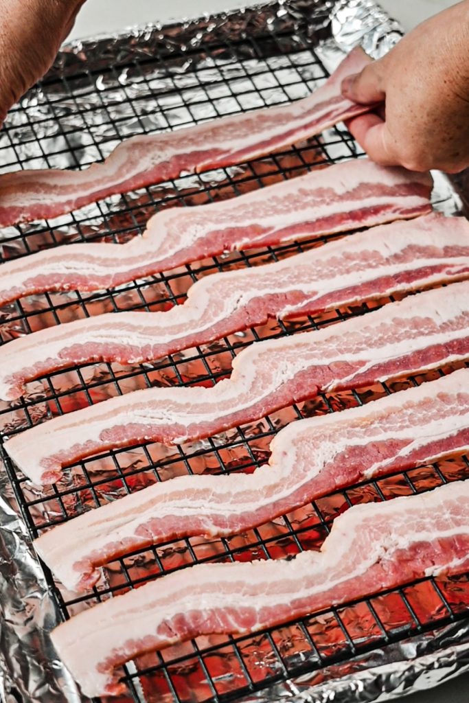 Raw bacon being lined up on a baking sheet lined with a wire rack.