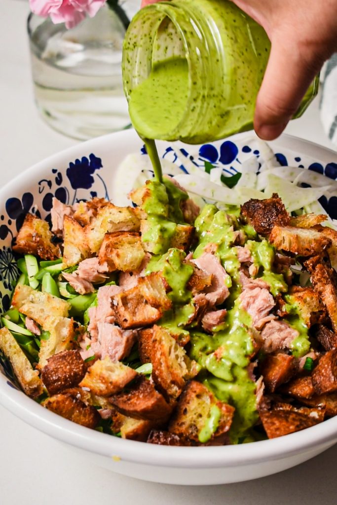 Pouring the pesto dressing over the raw asparagus white bean tuna salad in a bowl.